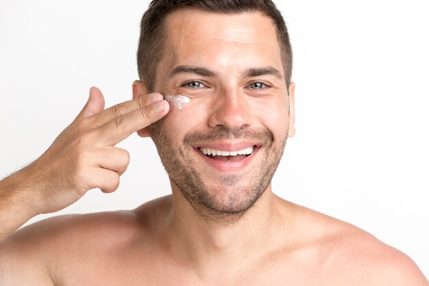 Smiling young man applying cream on face and looking at camera