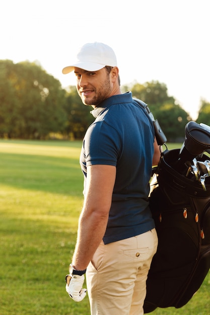 ゴルフバッグを保持しているキャップで若い男の笑みを浮かべてください。