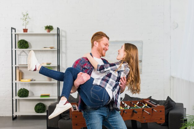 Smiling young man carrying her girlfriend in front of table soccer in the living room