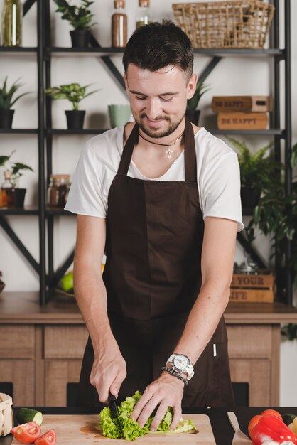 Smiling young man cutting lettuce with knife on chopping board