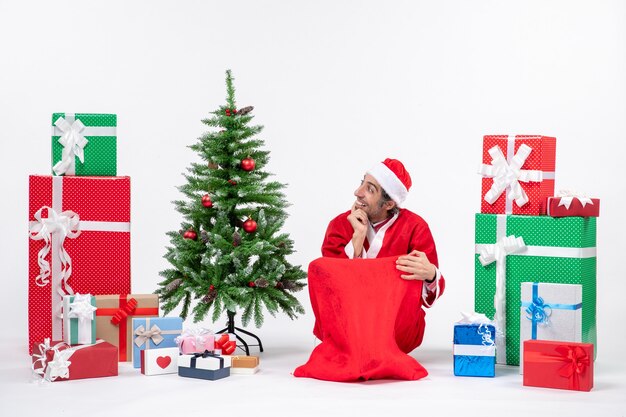 Smiling young man dressed as Santa claus with gifts and decorated Christmas tree sitting on the ground focused on something on the right side on white background