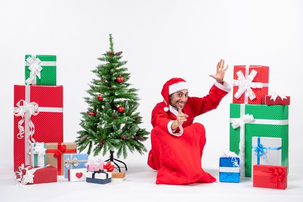 Улыбающийся молодой человек в костюме Санта-Клауса с подарками и украшенной елкой сидит на земле, указывая что-то вперед на белом фоне