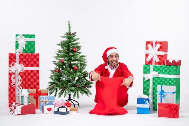 贈り物と白い背景の上の地面に座って飾られたクリスマスツリーとサンタクロースに扮した笑顔の若い男