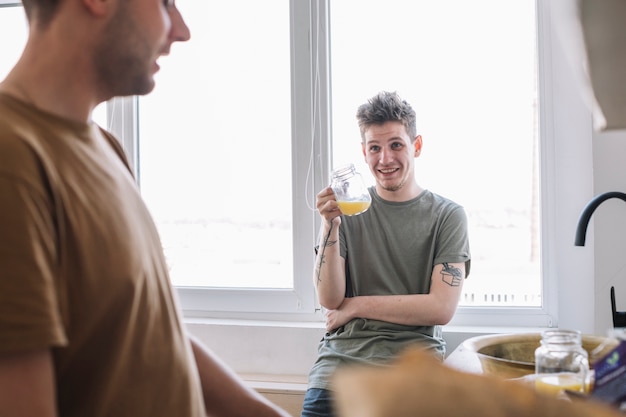 笑顔の若い男が台所で彼の友人を見てジュースを飲む
