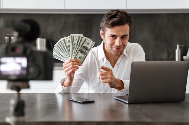 Free photo smiling young man filming his video blog episode about new tech devices while sitting at the kitchen table with laptop and showing bunch of money banknotes