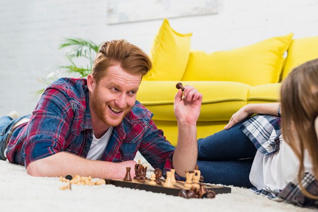 Smiling young man holding chess piece looking at her girlfriend lying on carpet