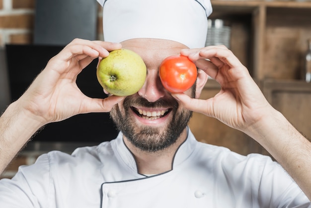 Smiling young man holding fresh ripe tomato and apple in front of his eyes