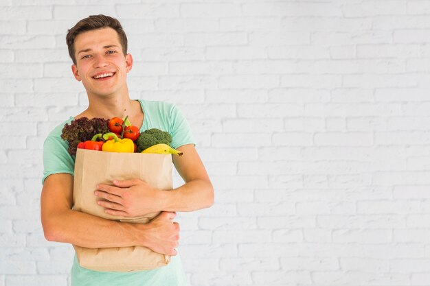 Smiling young man holding fresh vegetables and fruits in grocery paper bag