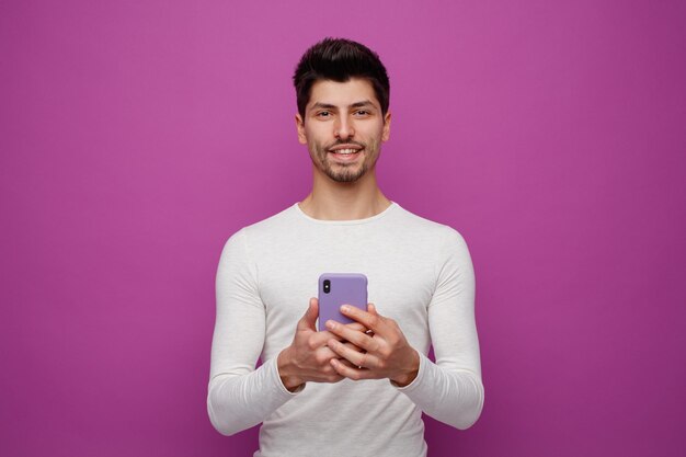 Smiling young man holding mobile phone looking at camera isolated on purple background