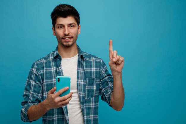コピースペースで青い背景に分離されたカメラを上向きに見ている携帯電話を保持している笑顔の若い男