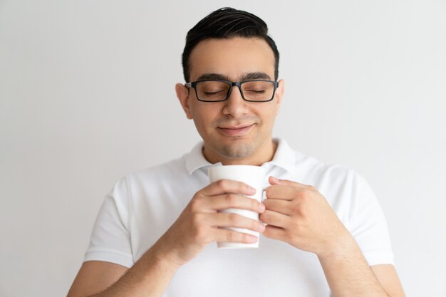 Smiling young man holding mug, smelling and drinking tea.