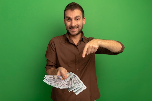 Smiling young man holding and pointing at money looking at front isolated on green wall