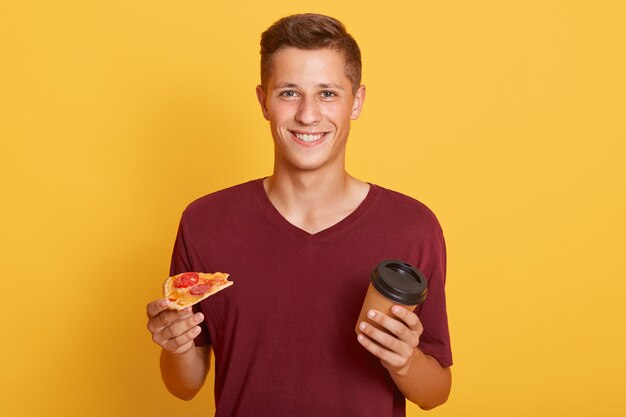 Smiling young man holding take away coffee and slice of tasty pizza,, having snack, wearing maroon t shirt