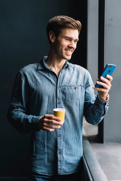 Улыбающийся молодой человек, холдинг выпить чашку кофе, глядя на мобильный телефон