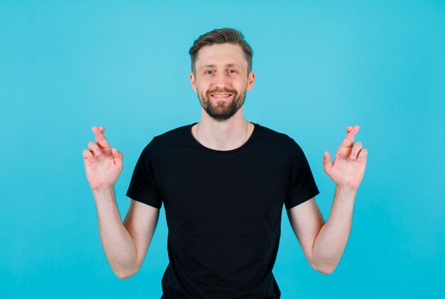 Smiling young man is looking at camera by crossing fingers on blue background