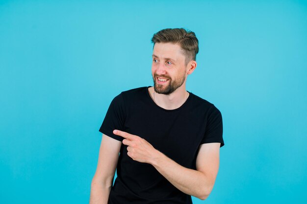 Smiling young man is looking left by pointing left with forefinger on blue background