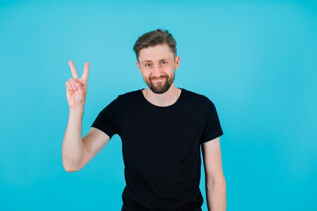 Smiling young man is showing victory gesture on blue background