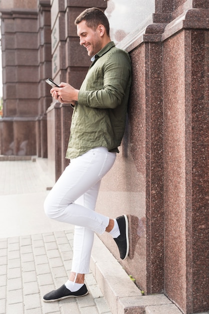 Smiling young man leaning on wall using cellphone