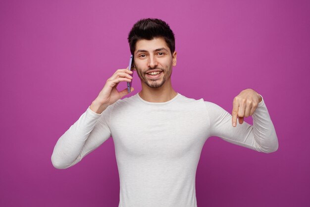 Smiling young man looking at camera pointing down while talking on phone isolated on purple background