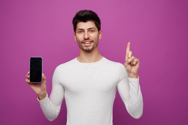 Smiling young man looking at camera pointing up showing mobile phone to camera isolated on purple background