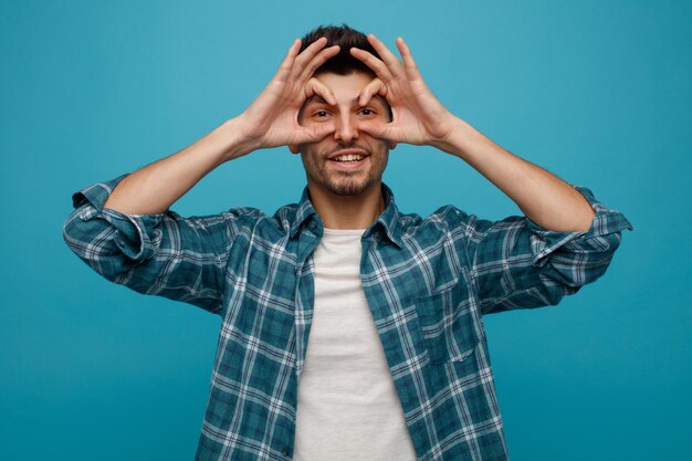 Smiling young man looking at camera showing binoculars gesture isolated on blue background