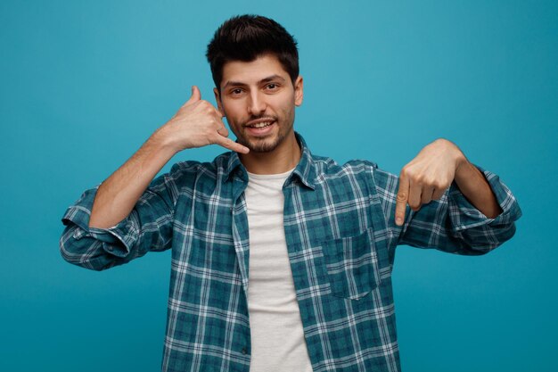 Smiling Young Man Looking At Camera Showing Call Me Gesture Pointing Down Isolated On Blue Background