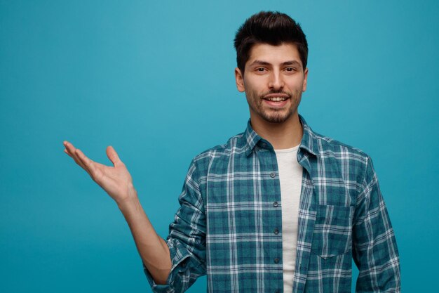 Smiling young man looking at camera showing empty hand isolated on blue background