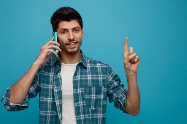 Smiling young man looking at camera talking on phone pointing up isolated on blue background