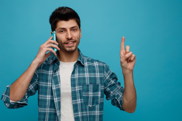 Smiling young man looking at camera talking on phone pointing up isolated on blue background