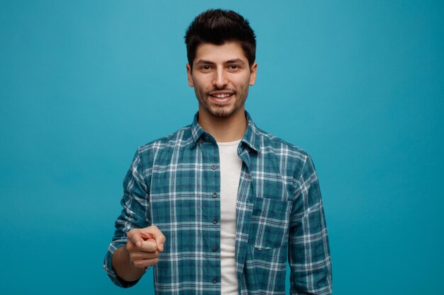 Smiling young man looking and pointing at camera isolated on blue background