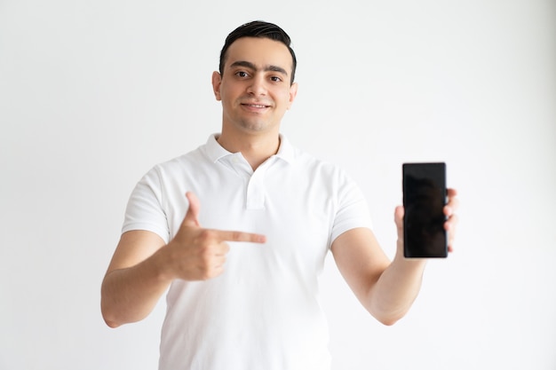 Smiling young man pointing at smart phone and looking at camera.