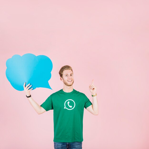 Smiling young man pointing upwards holding empty blue speech bubble