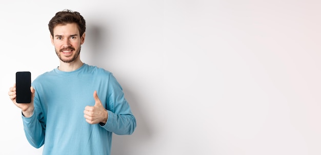 Smiling young man showing empty smartphone screen and thumbs up praising good application recommend