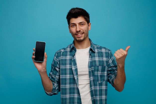 Smiling young man showing mobile phone to camera looking at camera pointing to side isolated on blue background