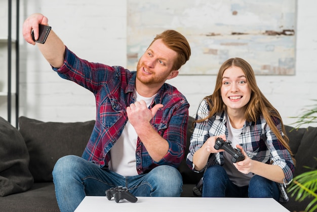 Foto gratuita giovane sorridente che mostra pollice sul segno mentre prendendo selfie sul telefono cellulare a casa