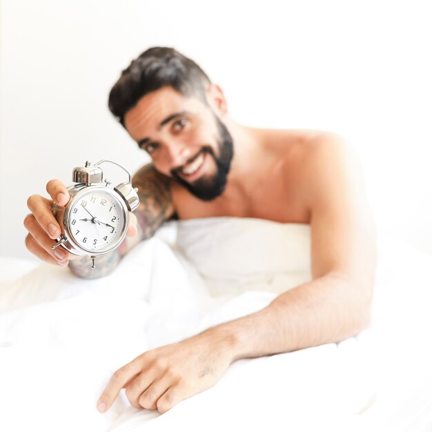 Smiling young man sitting on bed showing alarm clock