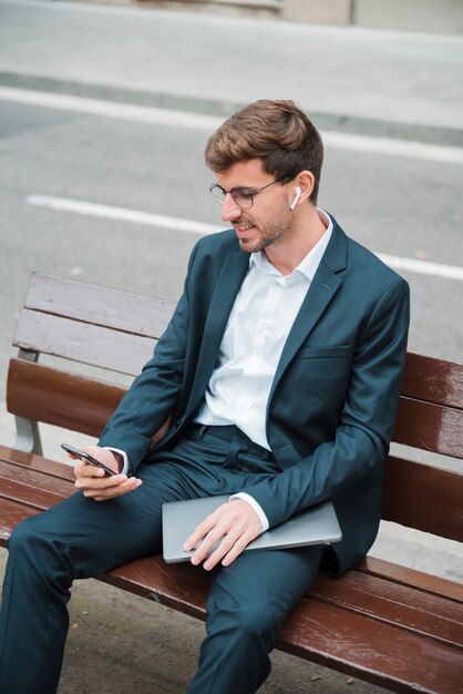 Smiling young man sitting on bench over the street using mobile phone with wireless earphone