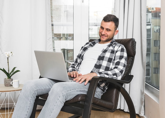 Улыбающийся молодой человек, сидя на стуле у себя дома с помощью мобильного телефона