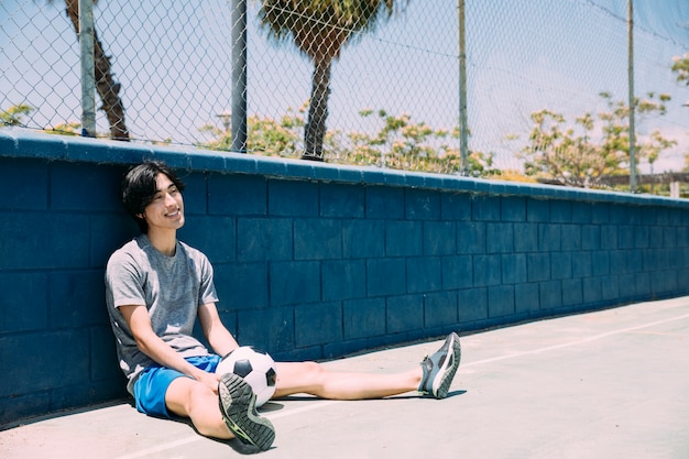 Free photo smiling young man sitting at fence with football