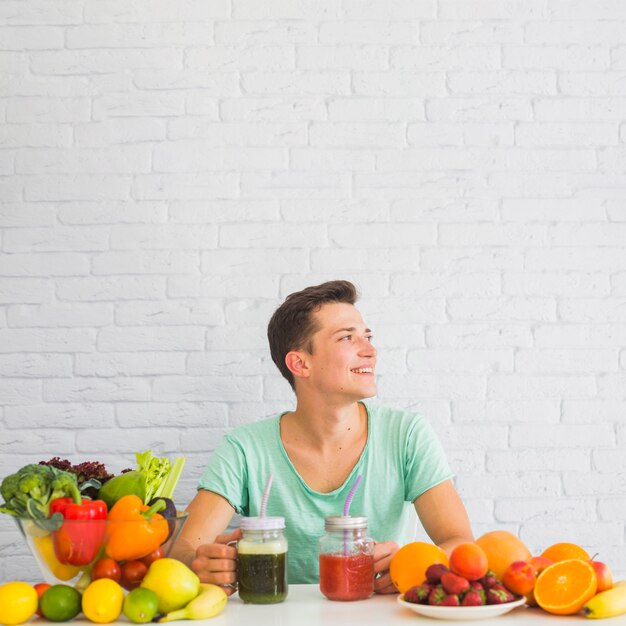 熟した、新鮮な野菜や果物のテーブルの後ろに座っている若い男を笑顔