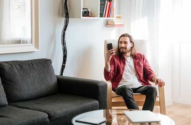 Smiling young man sitting and using mobile phone at home