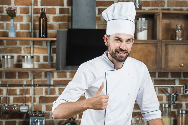Улыбаясь молодой человек, стоя на кухне, показывая пальцем вверх знак