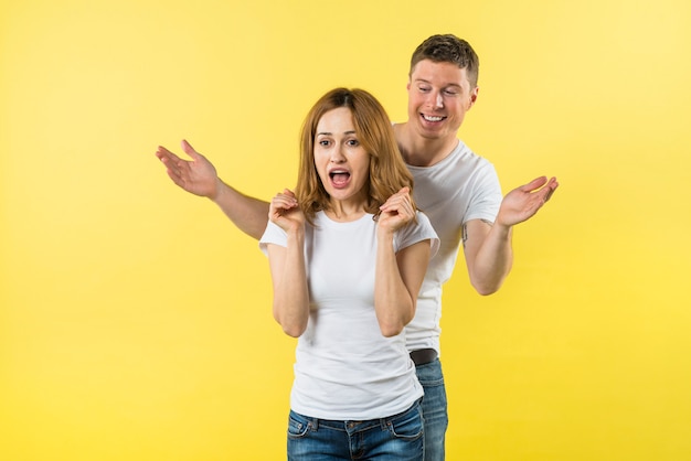 Free photo smiling young man standing behind the shocked girlfriend against yellow background