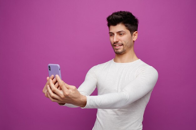 Улыбающийся молодой человек растягивает мобильный телефон, глядя на него на фиолетовом фоне