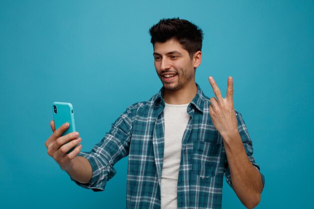 青い背景に分離されたビデオ通話を介して話している平和の兆候を示しているそれを見て携帯電話を伸ばして笑顔の若い男