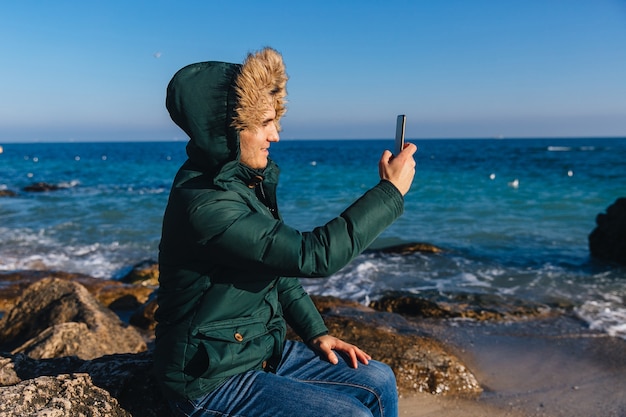 海の近くで携帯電話でセルフをしている若い男に笑顔。ファー付きの暖かいジャケットを着て
