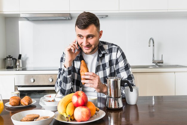 Smiling young man talking on mobile phone holding croissant in hand