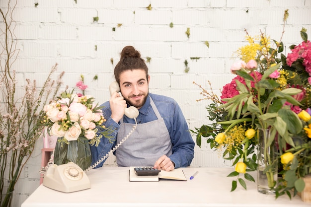 Улыбающийся молодой человек разговаривает по телефону с помощью калькулятора в цветочном магазине