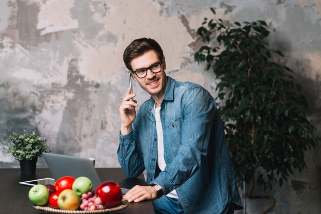 Улыбающийся молодой человек с помощью мобильного телефона с ноутбуком и фруктов