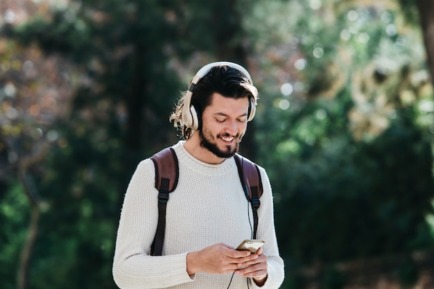 Улыбающийся молодой человек, используя телефон для прослушивания музыки на наушники в парке